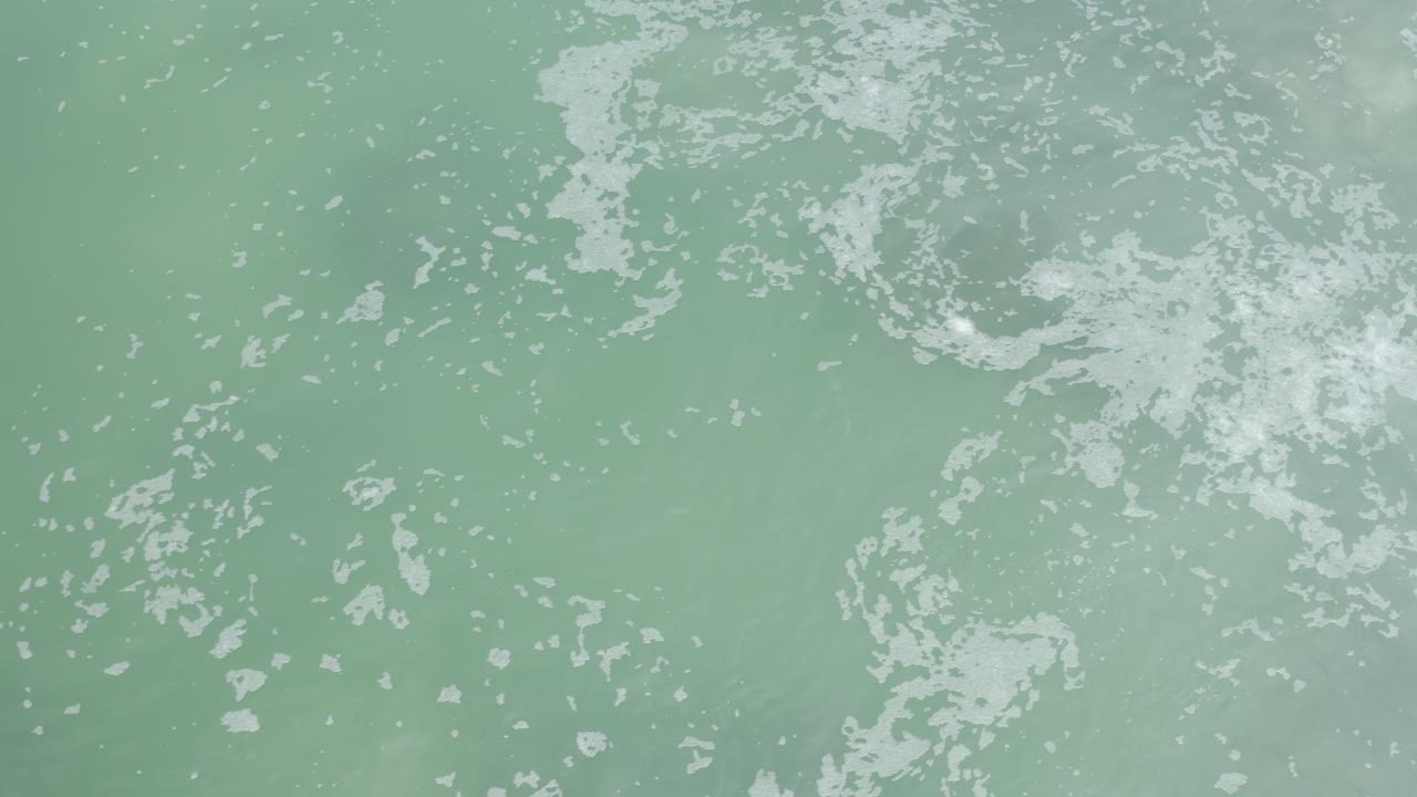 Overhead blue-green ocean water in Los Cabos with scattered white foam across the upper frame and no visible people.