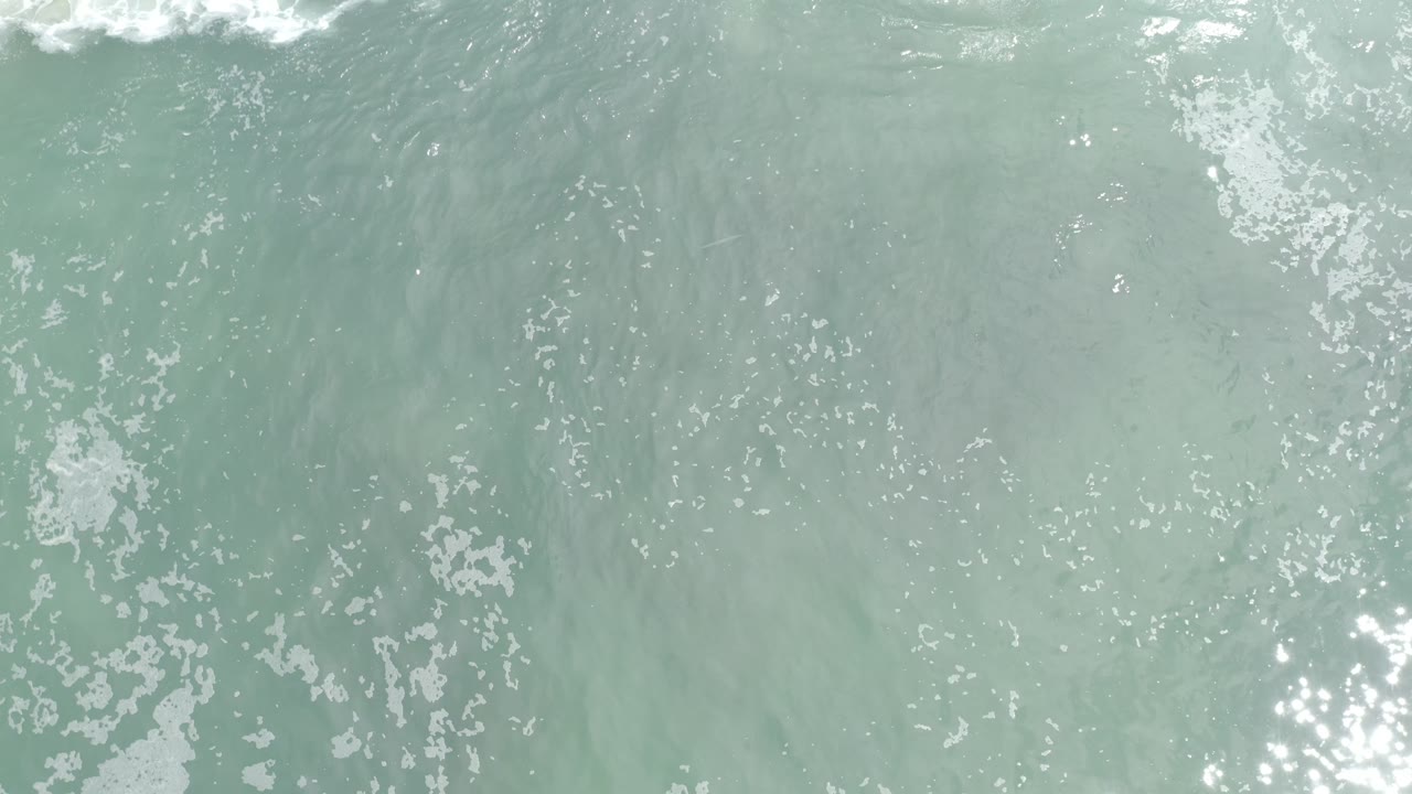 Overhead view of pale turquoise ocean water in Los Cabos with white foam near the upper edge, faint darker swirls below the surface, and no visible people.