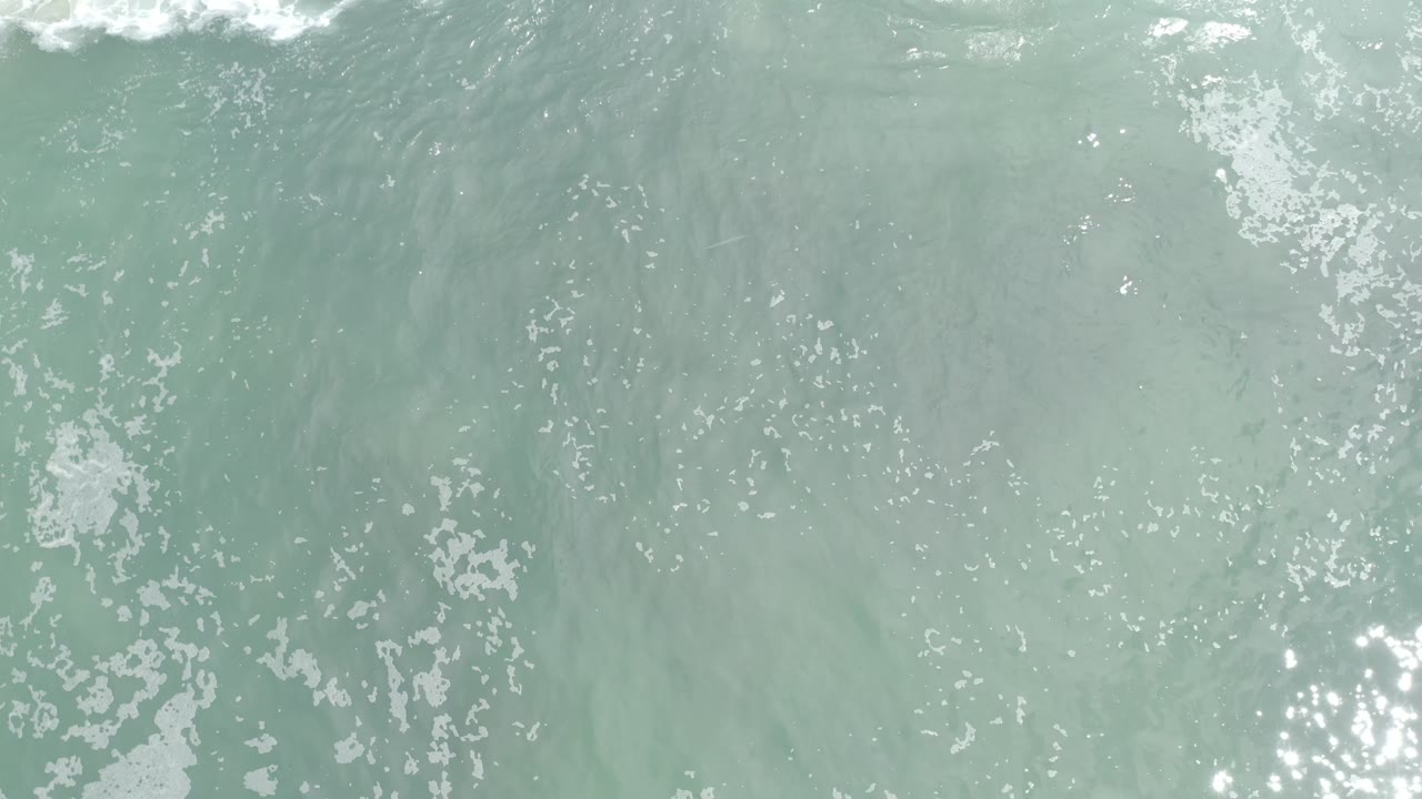 Aerial view over Los Cabos ocean water with scattered white foam across the left and center areas, pale green-blue color, and no visible people.