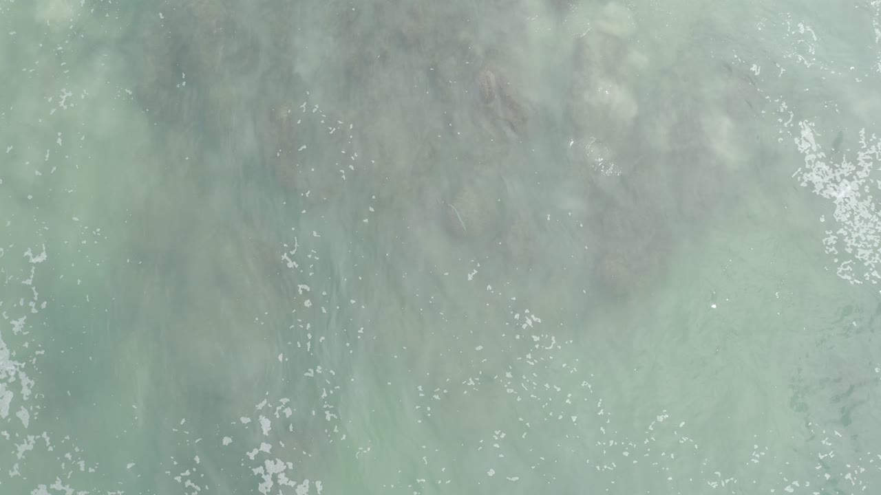 Overhead view of pale gray-green ocean water and scattered foam over shallow sand in Los Cabos, Mexico, with no visible people.