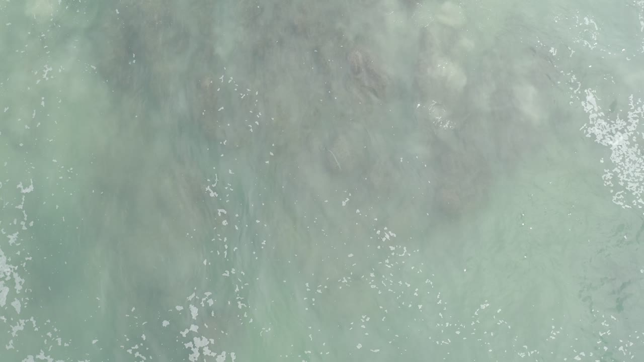 Overhead frame of soft gray-turquoise surf wash with fine white foam flecks crossing submerged sand in Los Cabos, Mexico, with no visible people.