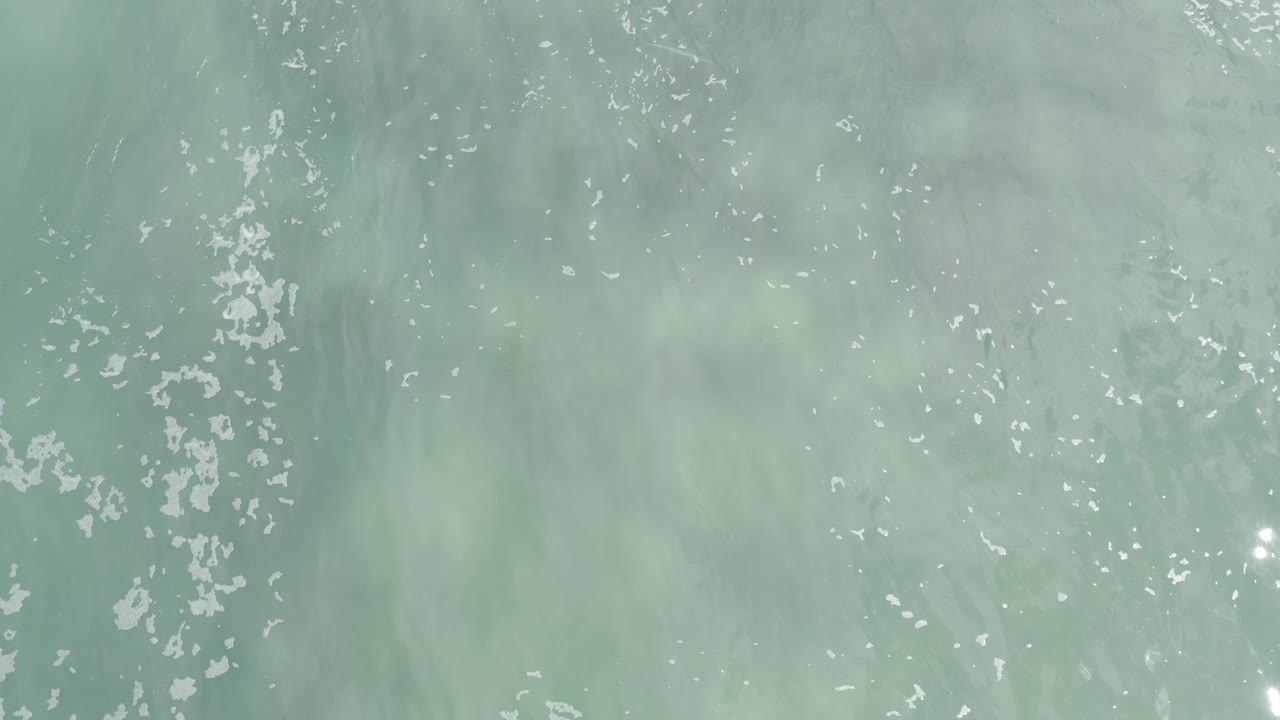 Overhead Los Cabos ocean water shows a vertical band of white foam slightly left of center, muted green-blue surf, and no visible people.