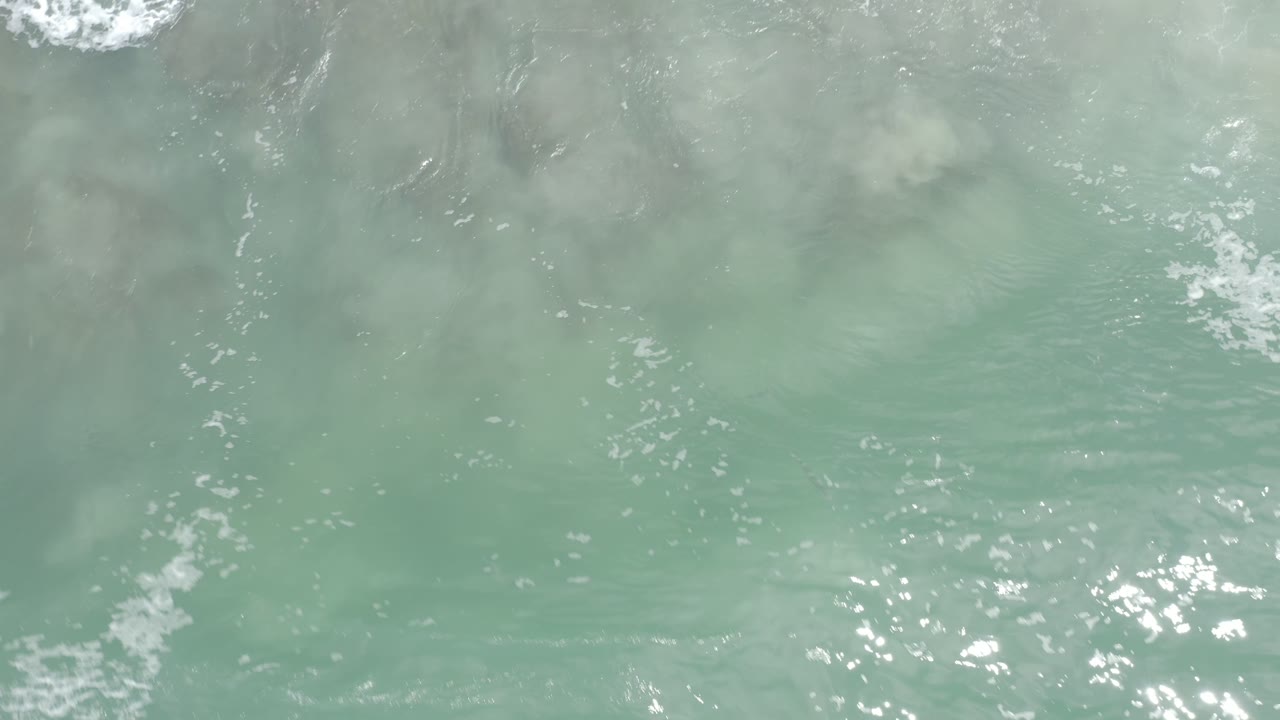 Top-down Los Cabos ocean water with a muted green-blue surface, cloudy sand streaks below, scattered sparkle, and no visible people.