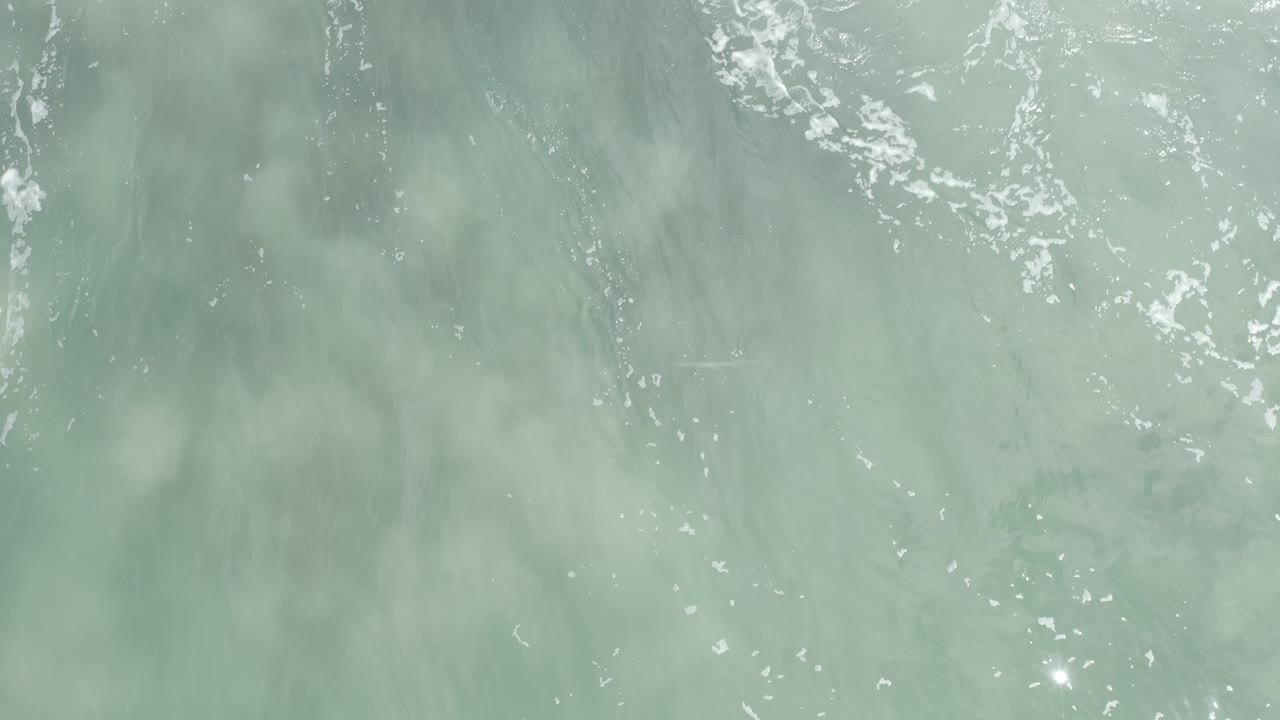 Top-down Los Cabos ocean surface with broken white foam near the top center, muted green-gray water below, and no visible people.
