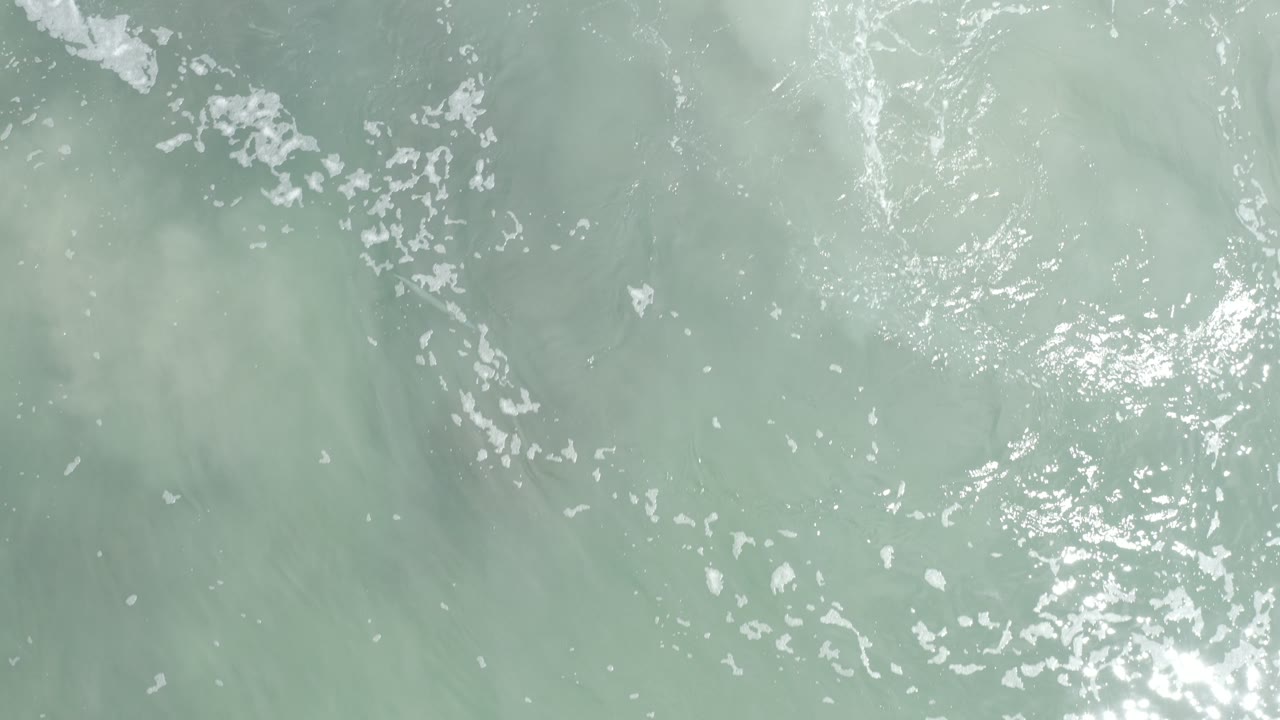 Top-down aerial view of pale turquoise ocean water in Los Cabos, Mexico, with scattered white foam and no visible people.