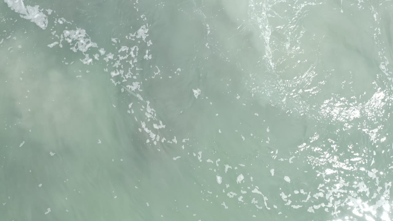 Aerial frame over pale green-blue water in Los Cabos, Mexico, with a brighter sun glint near the center and no visible people.