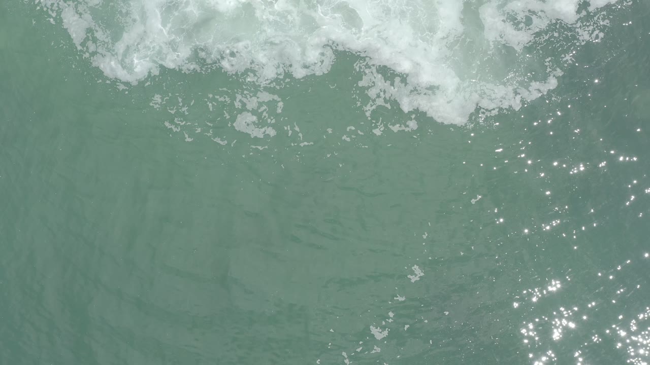 Overhead view of pale green seawater in Los Cabos, Mexico, with a band of white foam near the top and small sun glints on the surface, no visible people.