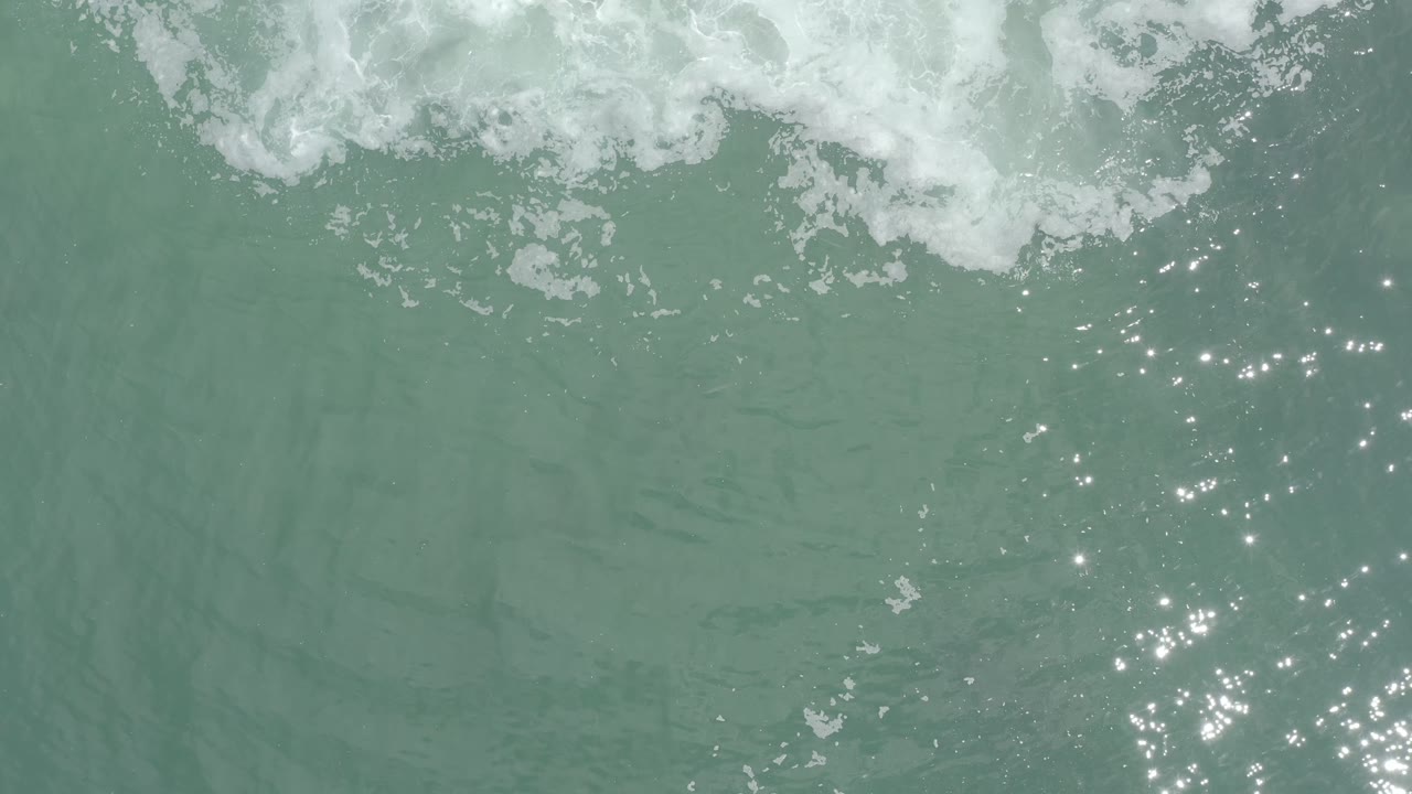 Overhead Los Cabos seawater with white foam along the upper edge and a cluster of bright sun reflections toward the lower right, no visible people.