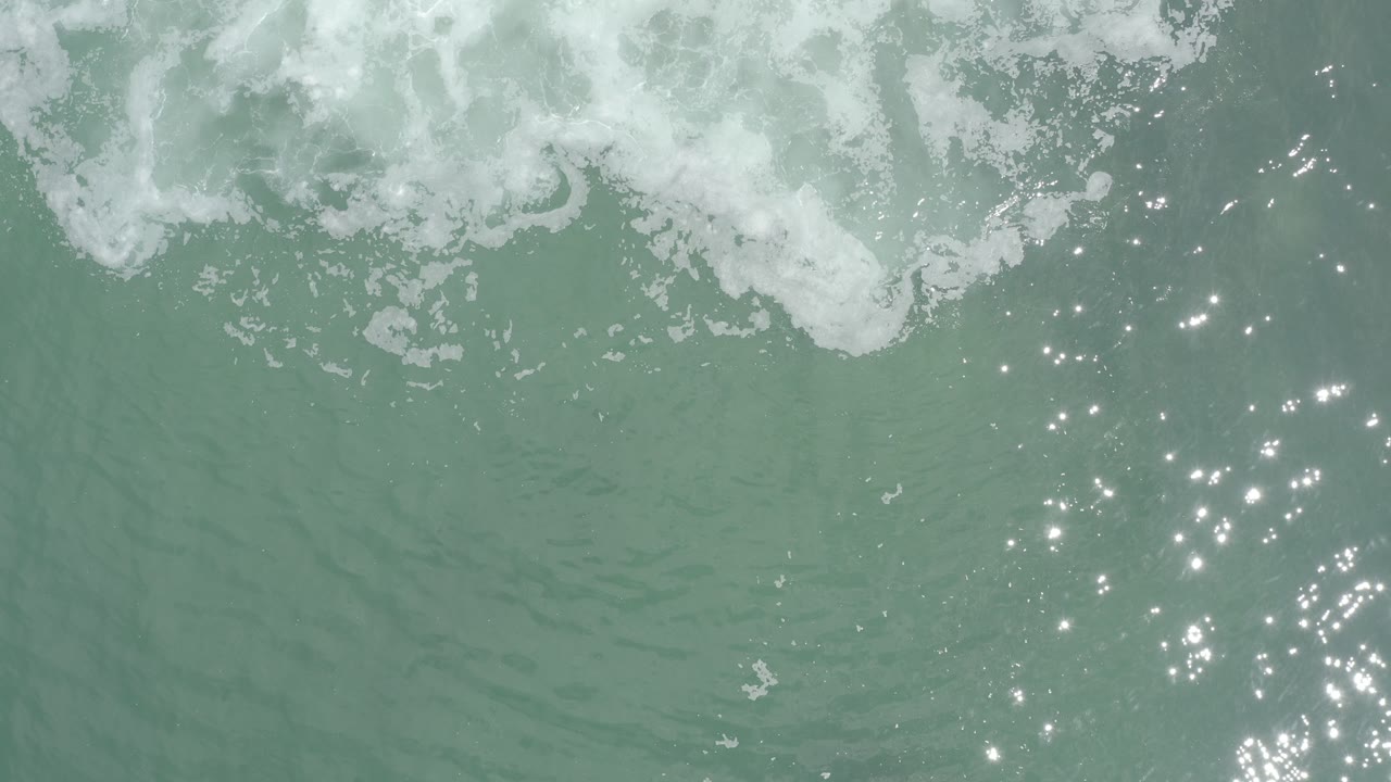 Overhead view of turquoise surf in Los Cabos, Mexico, with white foam along the top edge, scattered sun glints on rippling water, and no visible people.