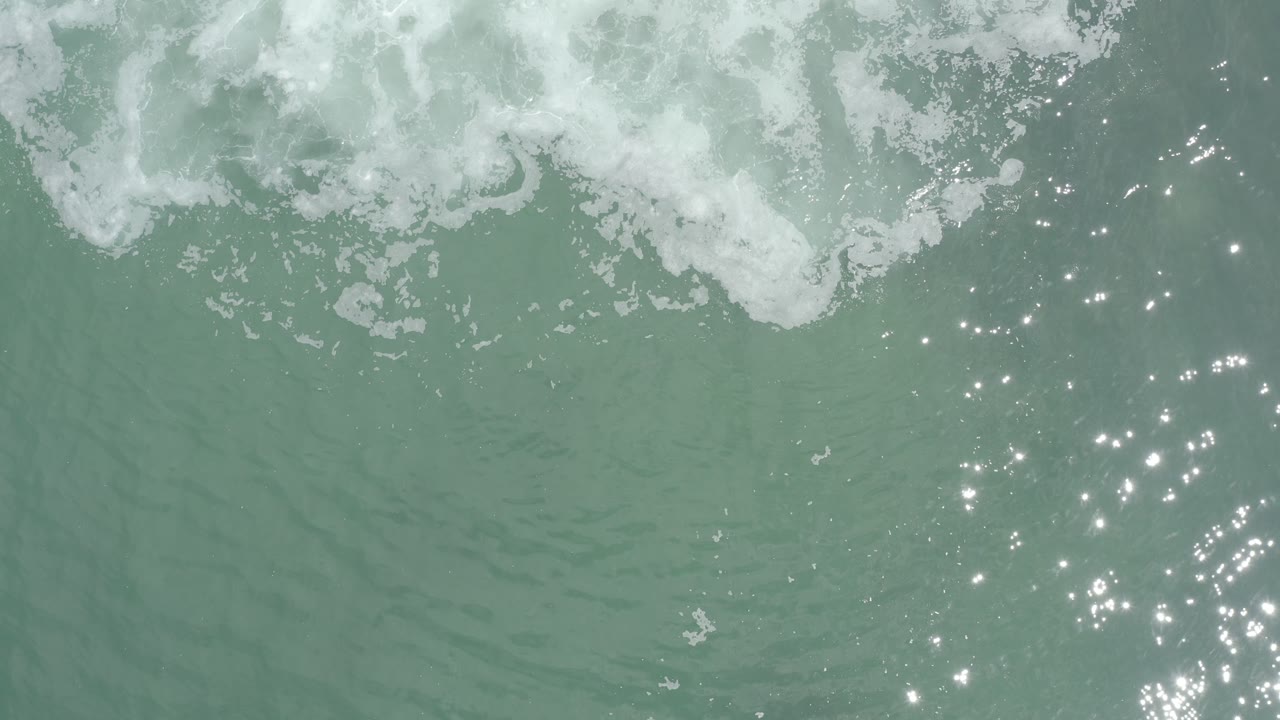 Foamy green surf in Los Cabos, Mexico, spreads across the upper frame while a dense patch of sunlight reflects from the rippled water below, with no visible people.
