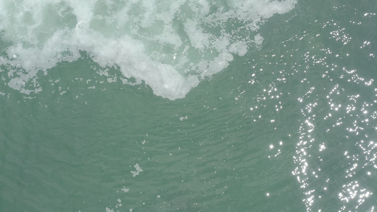 Overhead view of blue-green water in Los Cabos, Mexico, with white foam along the top edge, scattered sun reflections, and no visible people.