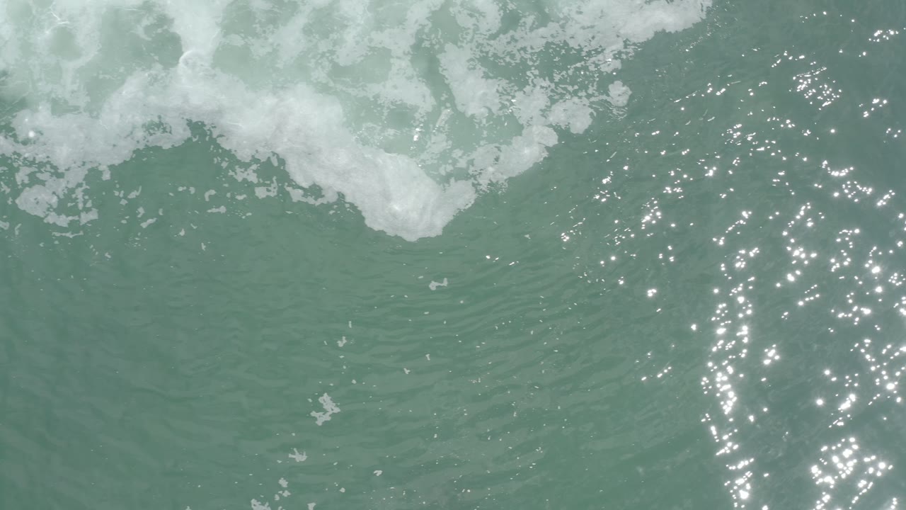Overhead Los Cabos water frame with a foamy band near the top, clustered sun sparkle near the center, soft ripples, and no visible people.