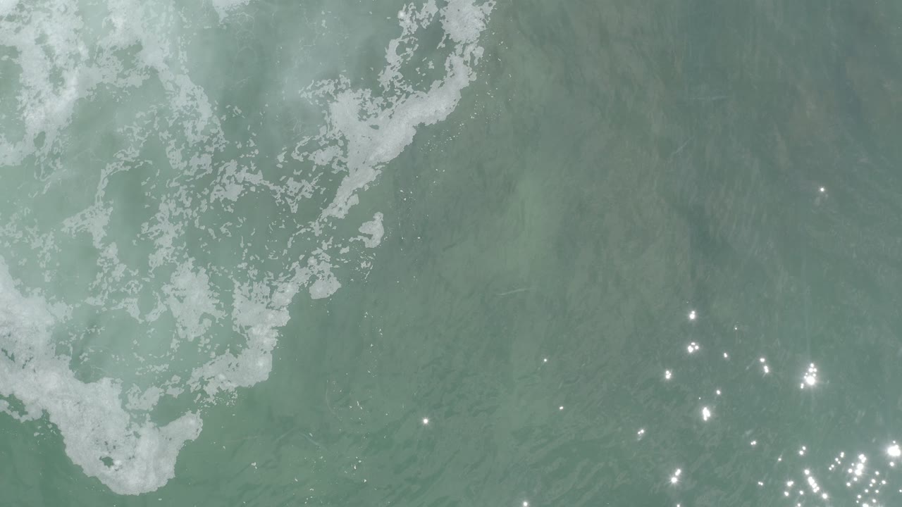 Opening overhead frame of Los Cabos ocean water with pale foam along the upper left and bright sun reflections near the lower right, with no visible people.