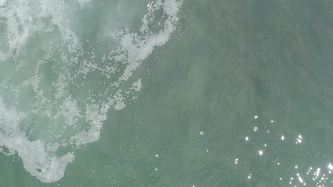 Overhead Los Cabos water frame with a white foamy wave edge near the upper center and a cluster of sun sparkles lower in the frame, with no visible people.