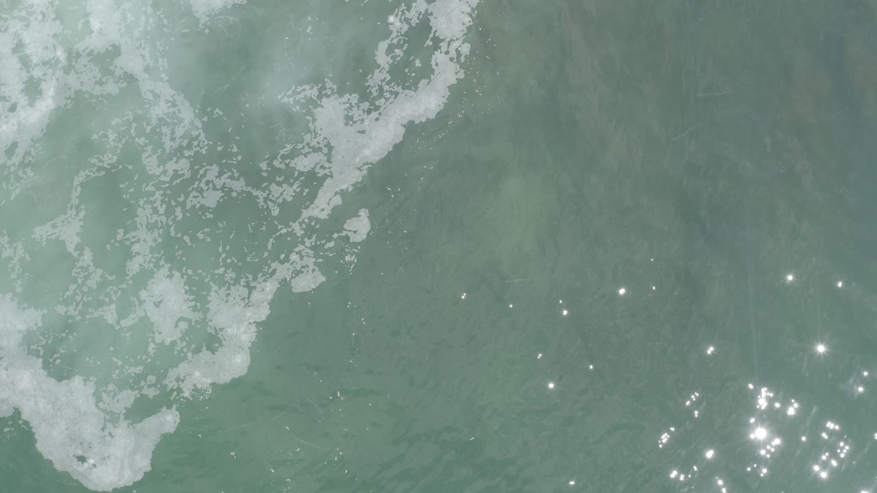 Aerial view of rippled ocean with foam and bright light flecks.