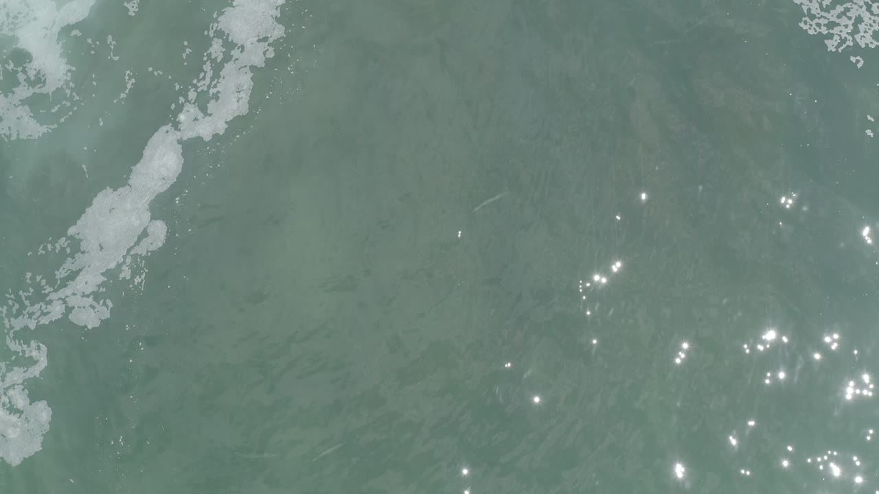 Turquoise water in Los Cabos fills the frame, with a broken white foam streak left of center and small bright reflections scattered across the lower middle, with no visible people.