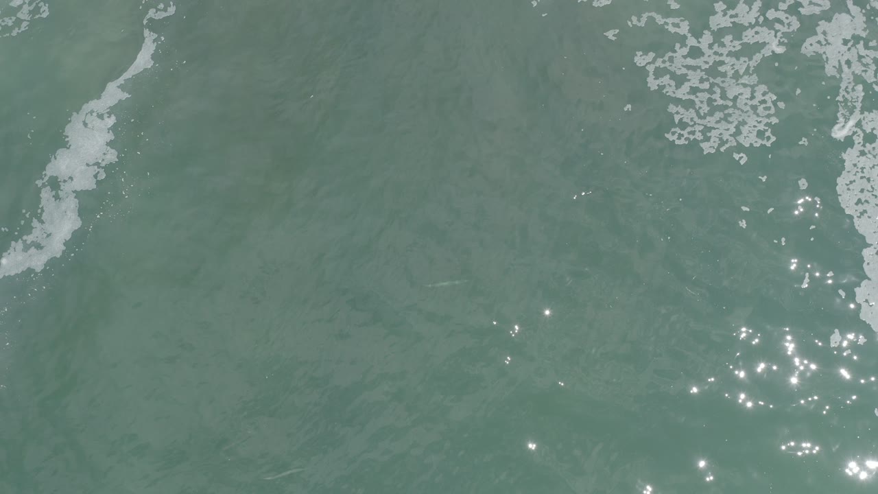 Overhead view of blue-green ocean water in Los Cabos, Mexico, with white foam entering from the upper left and scattered sun glints, no visible people.