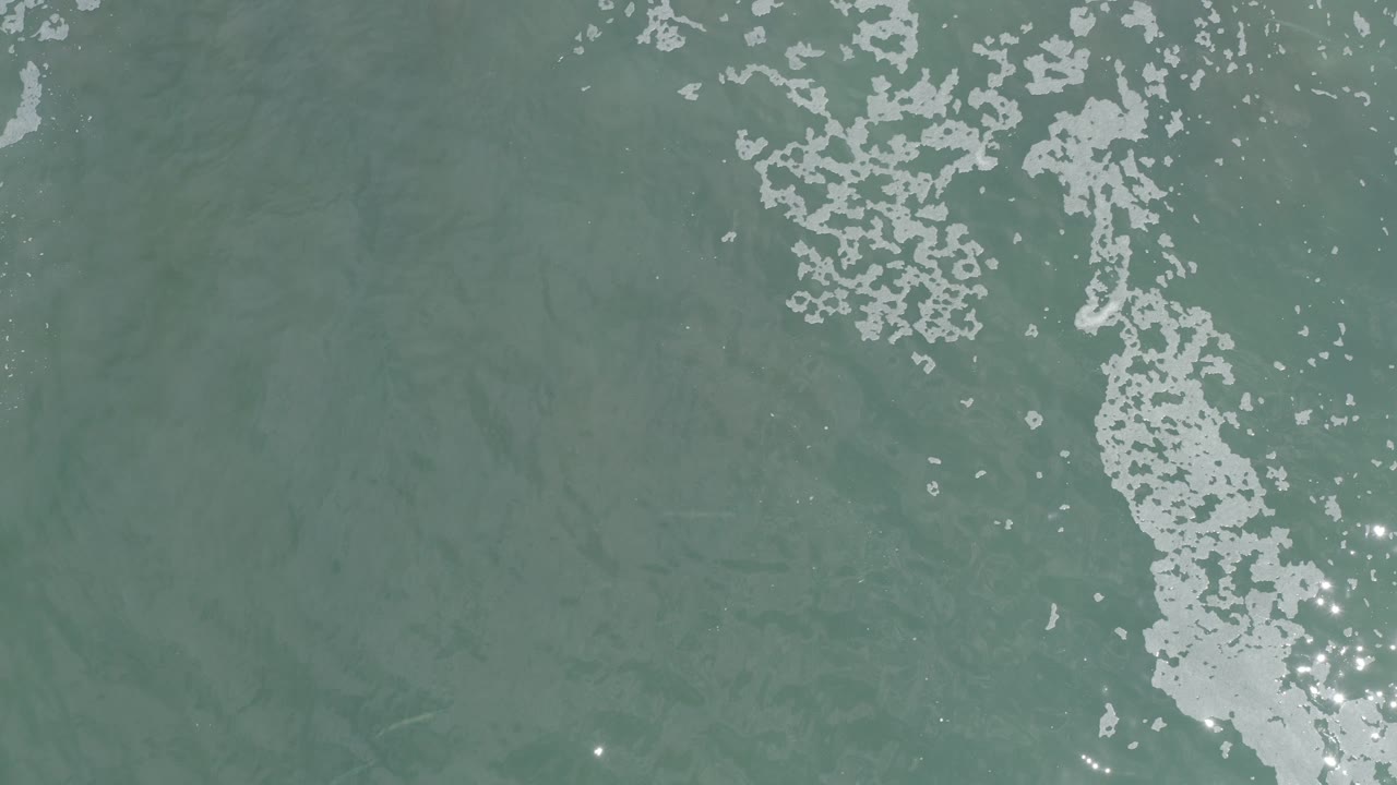 Overhead view of gray-green ocean water in Los Cabos, Mexico, with white foam streaking down the frame and no visible people.