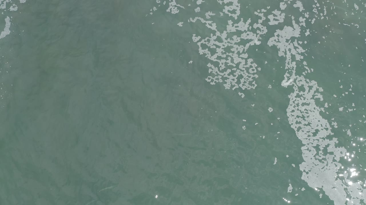 Aerial-looking view of Los Cabos ocean water with a vertical band of white foam and small bright reflections on the surface, no visible people.