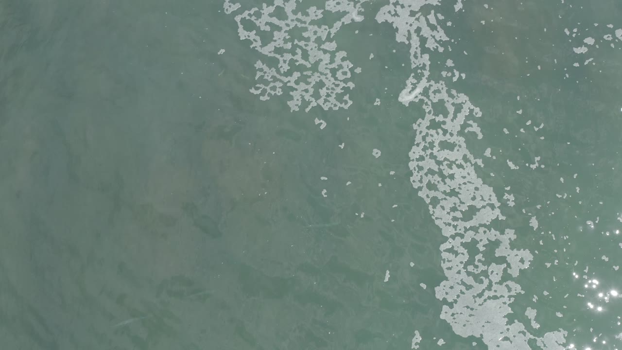 Overhead Los Cabos ocean frame with broken white foam running downward through the center and small sun glints on the right, with no visible people.