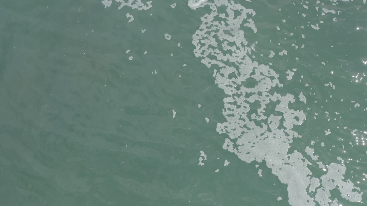 Overhead view of green-blue ocean water in Los Cabos, Mexico, with a vertical streak of white foam near the center and small sun reflections, no visible people.