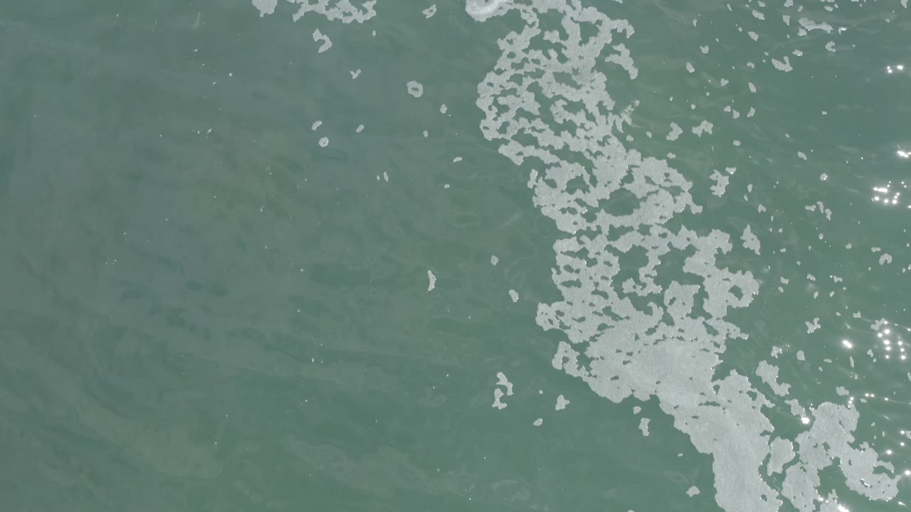 Overhead Los Cabos ocean surface with white foam running downward through the frame and clustered bright sun glints on the right, no visible people.