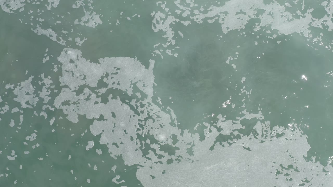 Top-down Los Cabos ocean surface with white foam broken into irregular islands across green-blue water and no visible people.