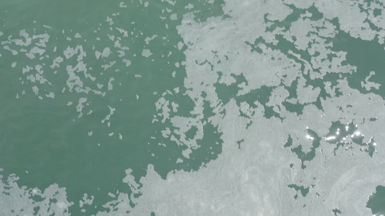 Overhead view of pale green sea water and patchy white foam in Los Cabos, Mexico, with no visible people.
