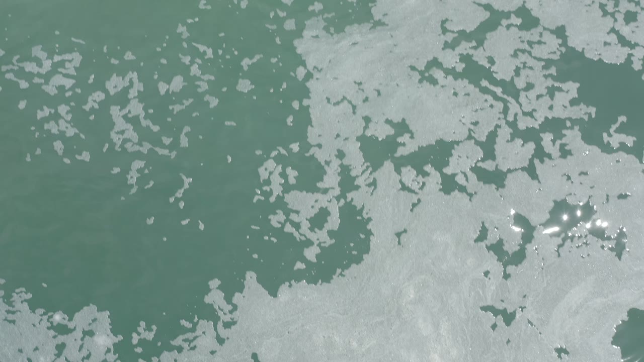 White foam spreads across green ocean water in an overhead Los Cabos frame, with small reflected highlights and no visible people.