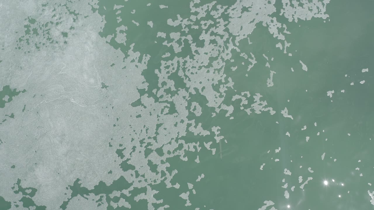 Overhead view of pale turquoise water in Los Cabos, Mexico, with broad white foam clusters at the left and upper edges and small sun glints near the lower center, no visible people.