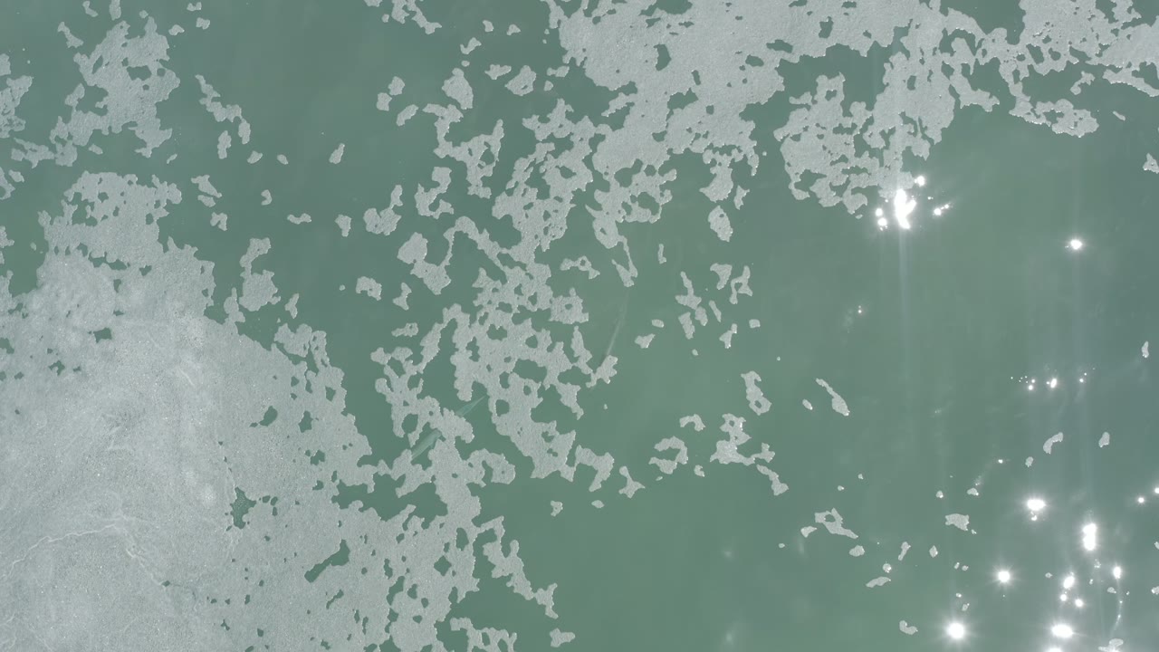 Overhead Los Cabos water surface with a broad white foam field across the top, smaller foam patches below, and no visible people.