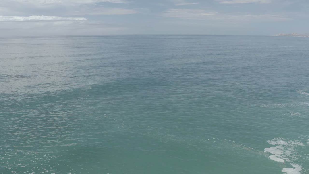 Opening aerial frame over the ocean near Los Cabos, Mexico, with turquoise water, a pale sky, faint foam at the lower edge, and no visible people.