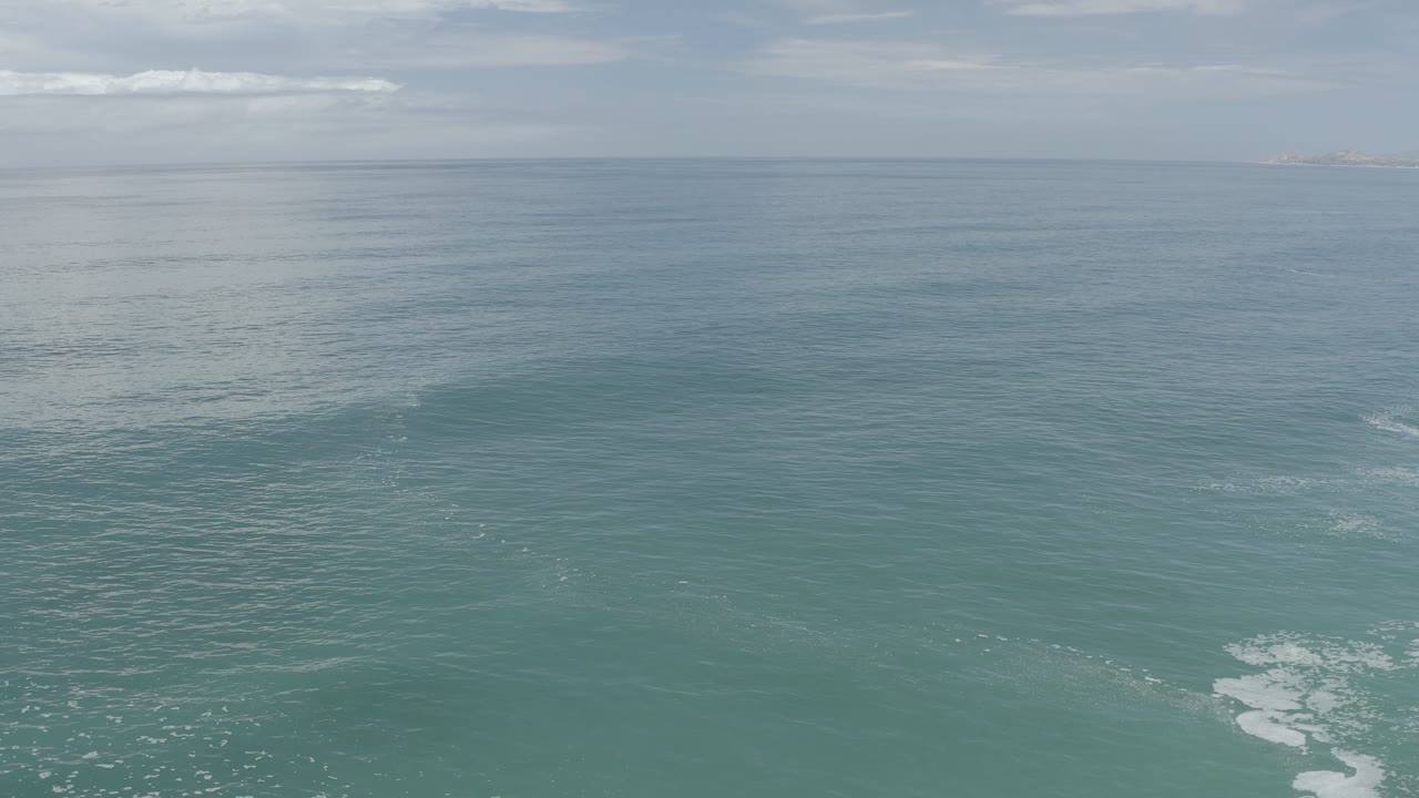 Aerial frame over Los Cabos ocean water with small white foam patches near the lower center, a level horizon, pale cloud cover, and no visible people.