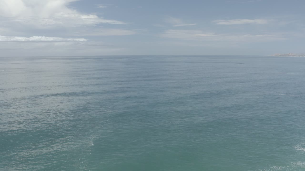Opening frame of calm blue-green ocean in Los Cabos, Mexico, with a level horizon, pale sky, bright cloud at upper left, faint distant land, and no visible people.