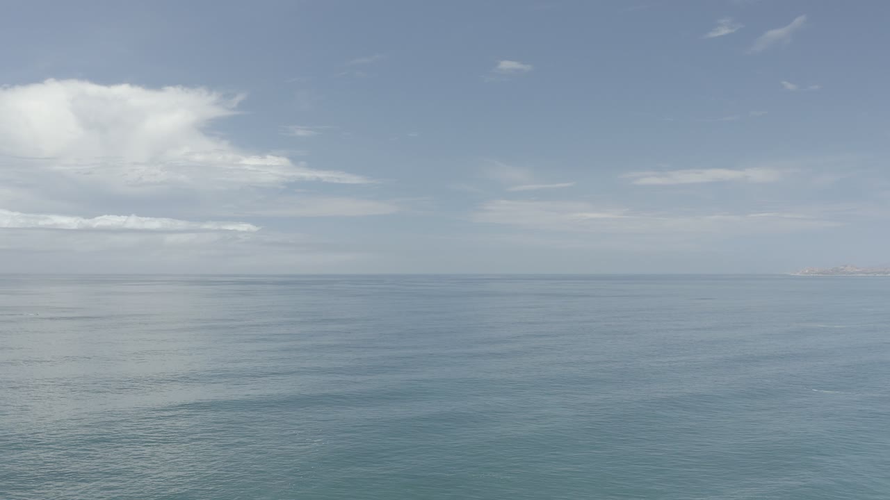 Wide aerial view over calm blue ocean in Los Cabos, Mexico, with a pale sky, scattered white clouds, a faint horizon line, and no visible people.