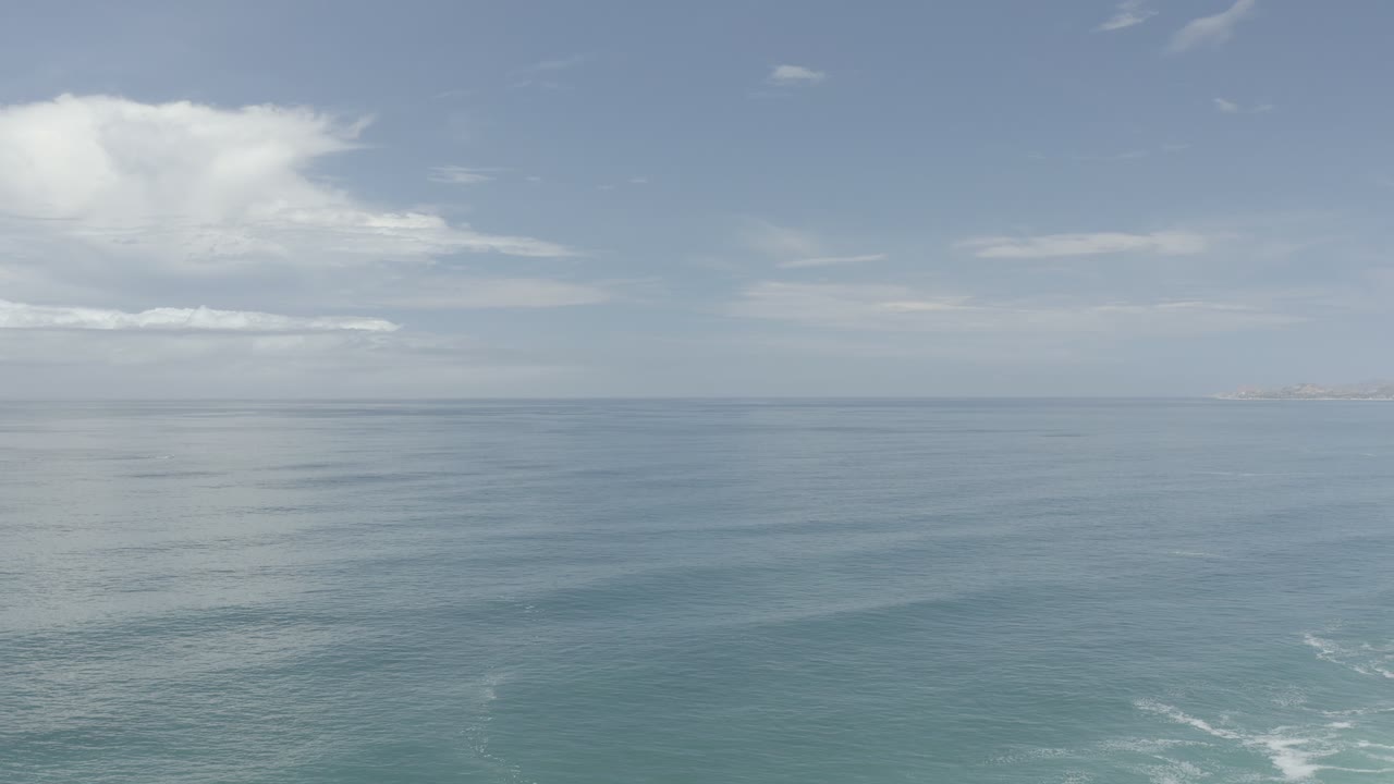 Los Cabos ocean frame with open blue-green water, a soft vertical cloud reflection, distant coastline near the horizon, and no visible people.