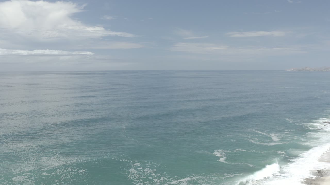 Opening frame of the Los Cabos coast with green-blue water in the foreground, a white surf line at the lower right, distant clouds, and no visible people.
