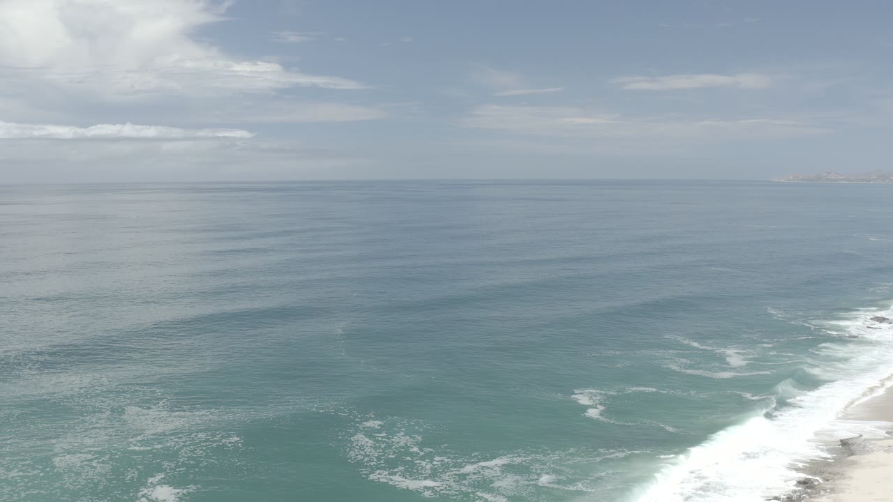 Open sea, white wave foam, beach edge, and thin clouds.