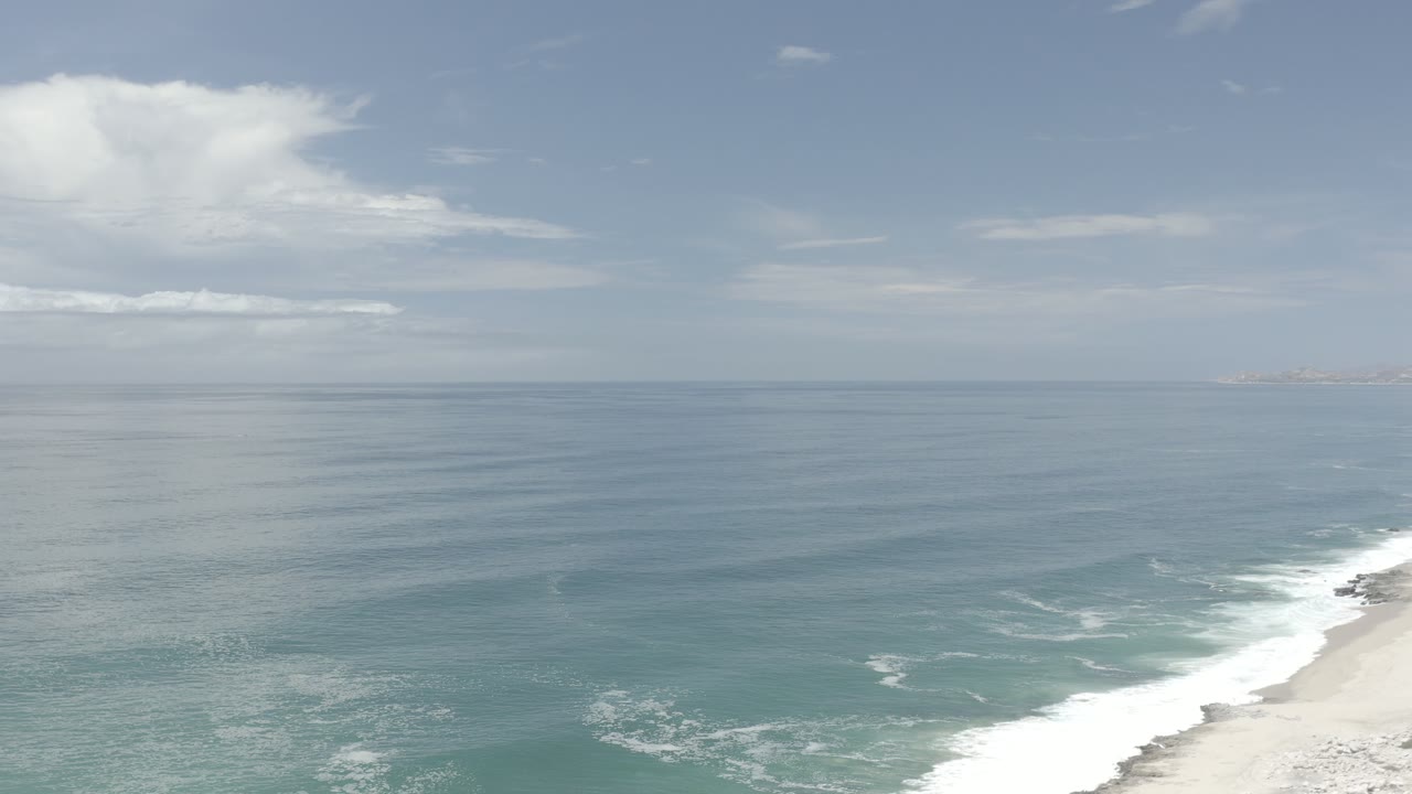 Early frame of the Los Cabos, Mexico shoreline with blue-green water, white surf at the lower right, a level horizon, scattered clouds, and no visible people.