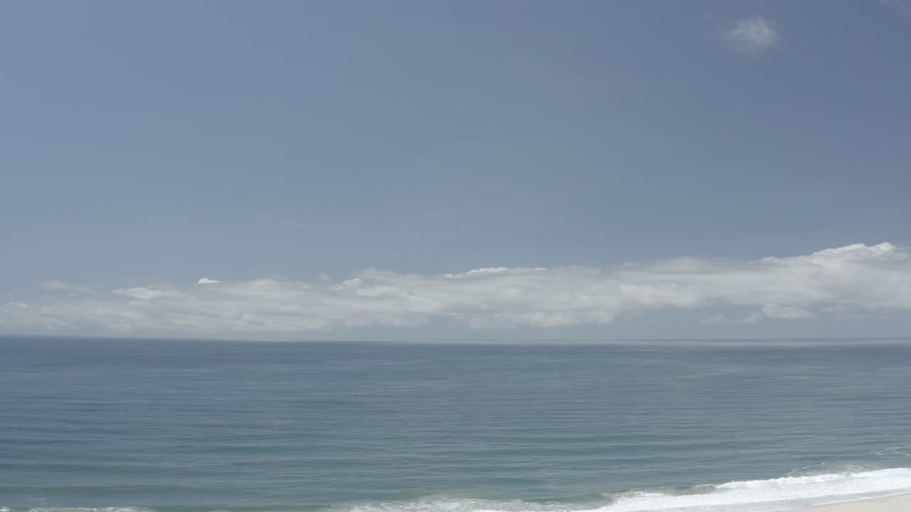 Opening frame of the Los Cabos ocean view, with a broad blue horizon, pale cloud band, white surf low in frame, and no visible people.