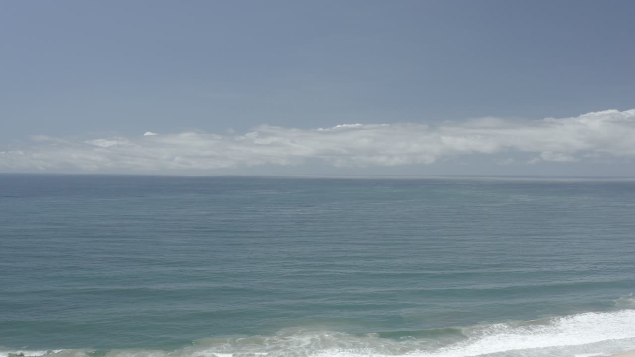 A Los Cabos, Mexico ocean view opens on blue-green water, a pale sky, low clouds, and a thin line of white surf at the bottom, with no visible people.