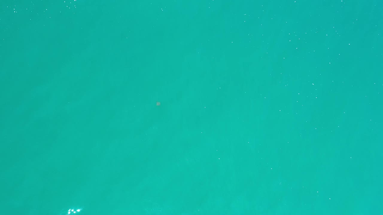 The Los Cabos water surface remains mostly even aqua, with faint ripples and a small cluster of reflected light near the lower area, no visible people.