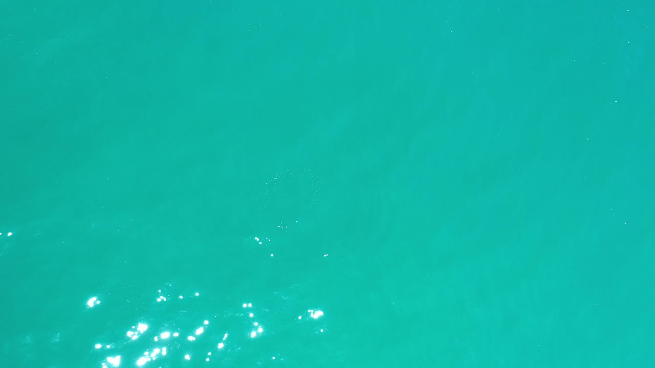 Los Cabos ocean water appears as a flat turquoise field with small clustered reflections and faint darker shapes below the surface, with no visible people.