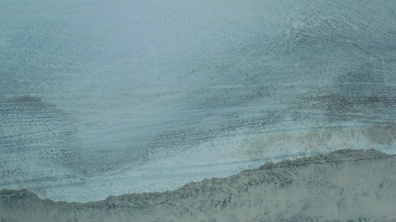 Overhead frame from Los Cabos, Mexico showing muted blue surf washing onto pale sand, with no visible people.