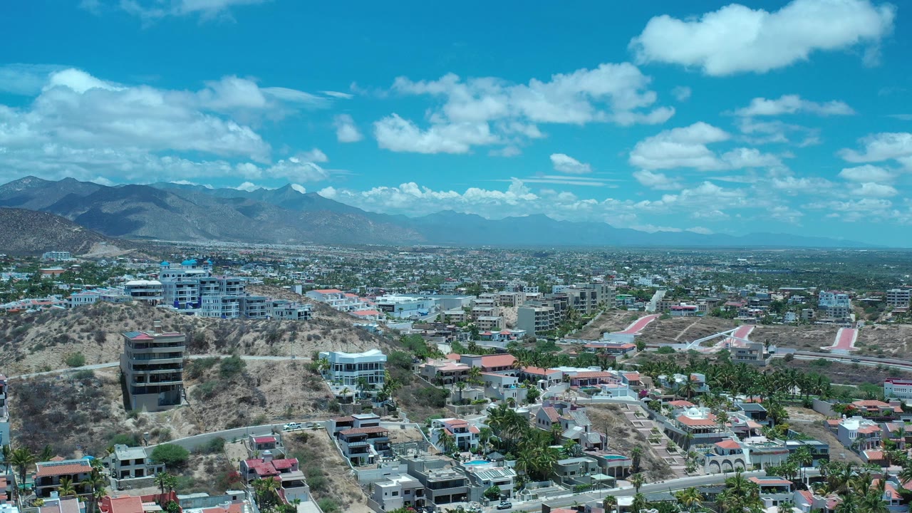 Aerial view of hillside buildings, dry slopes, distant mountains, and blue sky.