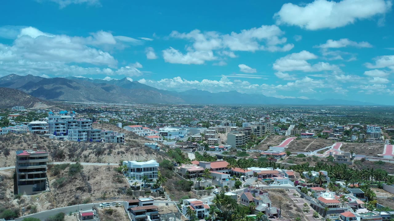 Los Cabos aerial view one second in, with a dark mid-rise building near the lower left, white hillside structures, broad urban sprawl, mountains, and no visible people.