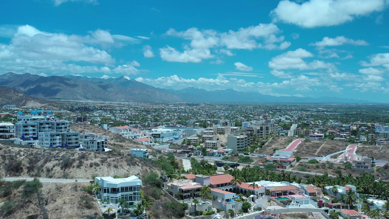 Early frame in Los Cabos, Mexico, showing more of the foreground ridge, clustered homes, a broad urban plain, mountains to the left, bright clouds, and no visible people.