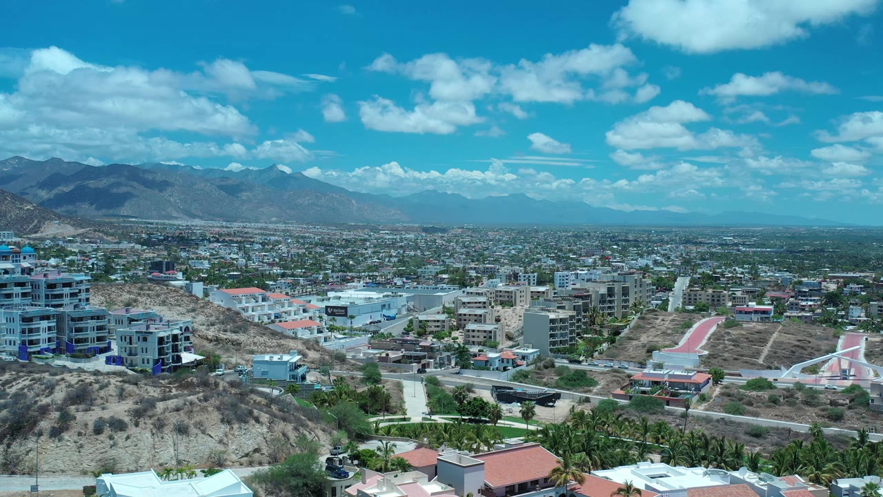 Early frame of the Los Cabos town view with a rocky foreground hill, clusters of low buildings, a broad mountain range, bright blue sky, and no visible people.
