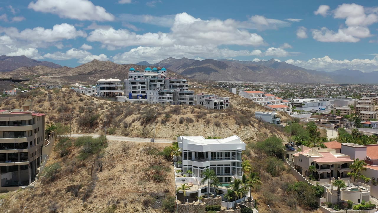 Elevated Los Cabos view with white houses on a dry slope, beige residential buildings nearby, distant urban development, and no visible people.