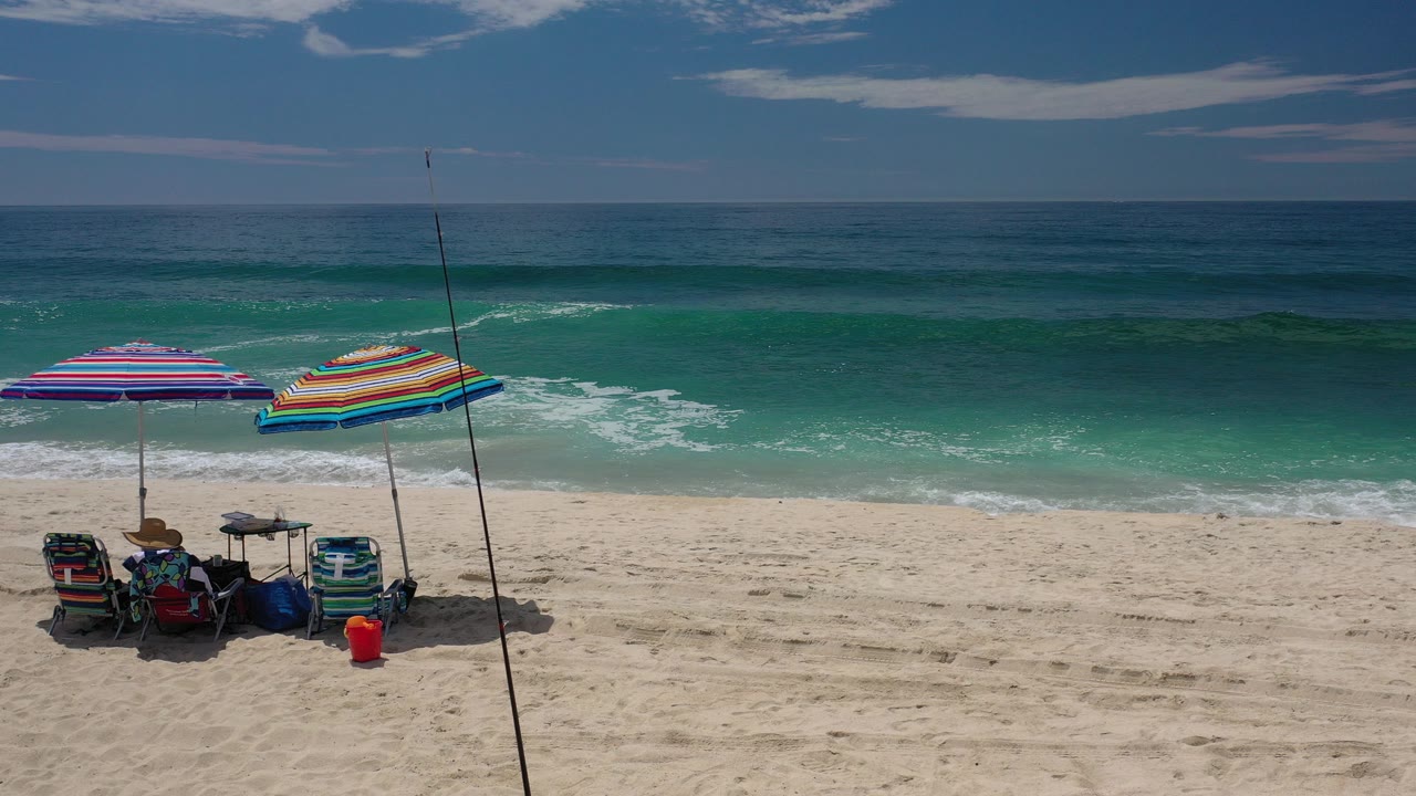 Opening frame of a Los Cabos beach with two striped umbrellas, folding chairs, bags, a red bucket, pale sand, turquoise water, and no visible people.